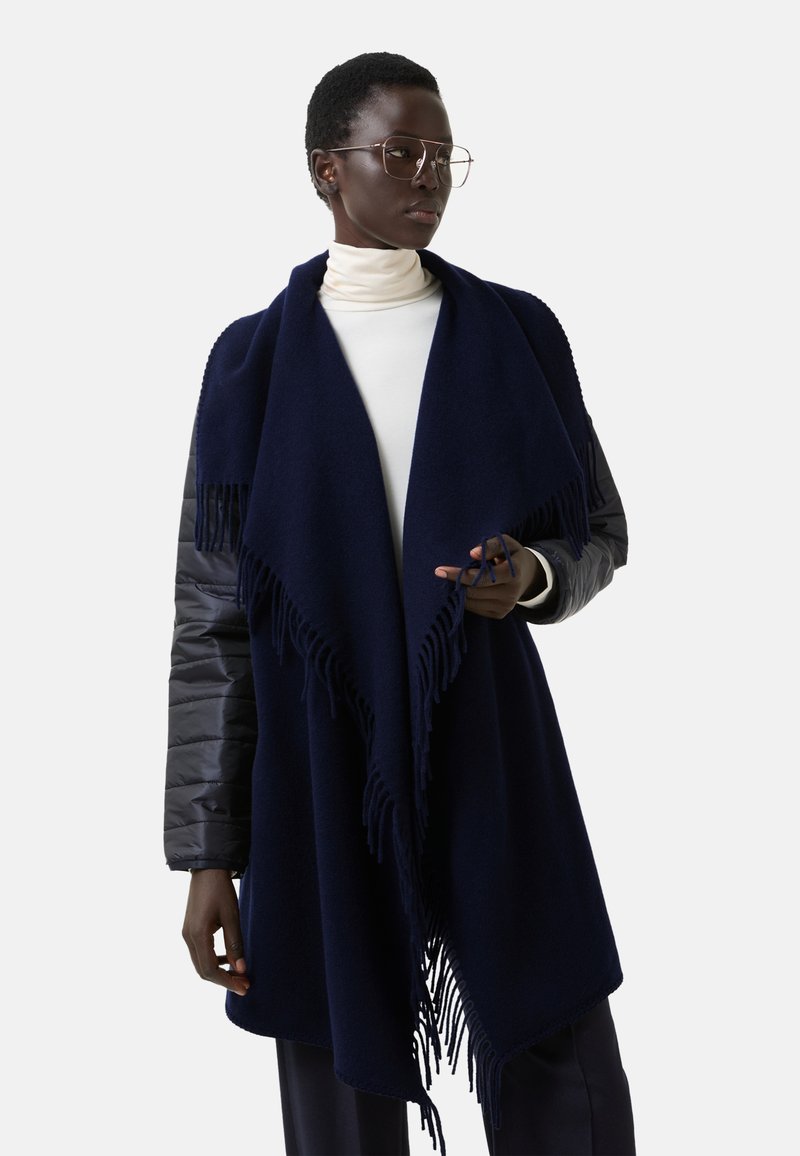 Námořnická modrá šála s třásněmi, vyrobená z měkkého materiálu; vrstvená přes světlý top s vysokým límcem, doplněná šedým rukávem.