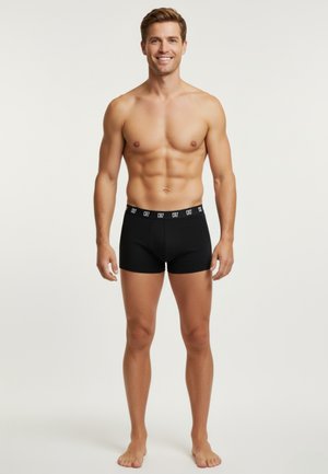Smilende muskuløs mand stående barfodet iført sorte boxer briefs med CR7-logo i taljebåndet på ensartet lys baggrund.