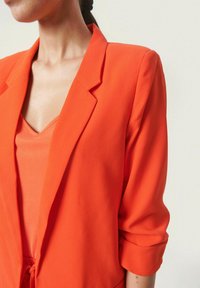 Pomarańczowa marynarka z wyrazistym kołnierzem i podwiniętymi rękawami, wykonana z gładkiego materiału. Noszona na pasującym pomarańczowym topie z dekoltem w serek.