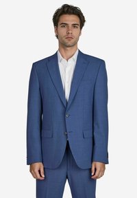 Ein Mann in einem maßgeschneiderten blauen Anzug steht vor einem wei�ßen Hintergrund, blickt direkt in die Kamera und hat einen ernsten Gesichtsausdruck.