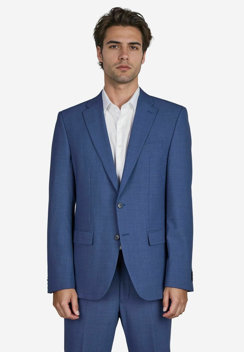 Ein Mann in einem maßgeschneiderten blauen Anzug steht vor einem weißen Hintergrund, blickt direkt in die Kamera und hat einen ernsten Gesichtsausdruck.