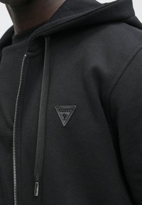 Sudadera con capucha negra con cremallera, tela texturizada, cordones estilo cordón de zapato y un logotipo triangular en relieve de "GUESS" en el pecho.