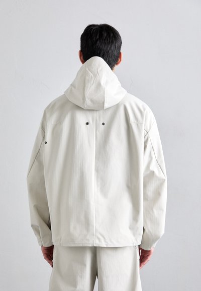 Chaqueta beige claro con capucha, fabricada en tejido suave. Presenta un corte holgado, hombros caídos y dos agujeros de ventilación en la parte posterior.