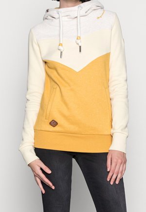 Kobieta ubrana w kapturówkę w kolorach białym i musztardowym z przednimi kieszeniami oraz czarne spodnie, stojąca na jednolitym tle.