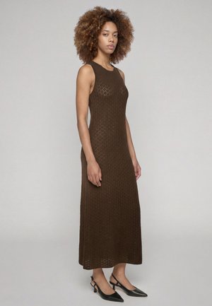 Donna con capelli ricci che indossa un abito marrone in maglia senza maniche lungo e tacchi bassi neri a punta, in piedi davanti a uno sfondo semplice.