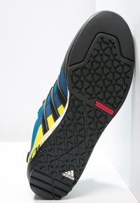 Blå sportskor med svarta och gula ränder. Gummisula med hexagonalt mönster för grepp. "TRAXION" och Adidas-logotyp i vitt.