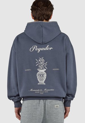 Osoba v tmavě modré mikině s nápisem „Pegador“ a bílým květinovým vzorem na zadní straně, doplněná o šedé texturované kalhoty.