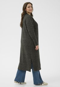 Temno siva, rebrasta pletena jopica s stranskimi razpoki, oblečena čez razširjene jeans hlače. Tekstura se zdi mehka in prijetna ter ima sproščen kroj.