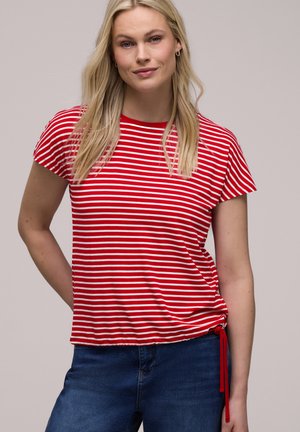 Vaalea nainen, joka pukeutunut puna-valkoisiin raidallisiin lyhythihaisiin puseroon, jossa on nyöritys, yhdistettynä sinisiin farkkuihin, yksiväristä taustaa vasten.