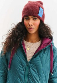 Kaštanová pletená čepice s modrým štítkem, zelená prošívaná bunda s kaštanovými doplňky, krémový svetr s copánkovým vzorem, zlatý přívěsek na náhrdelníku.