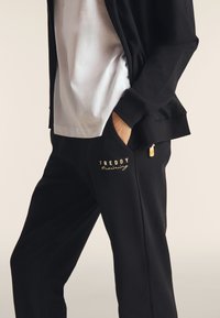 Pantalon de sport noir avec un logo "Freddy" doré, poches latérales et une texture lisse, associé à un T-shirt blanc et une veste zippée noire.