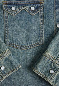 Sezione giacca di jeans con una tonalità blu chiaro, cuciture arancioni a contrasto, chiusure a pressione tipo perla e un design della tasca a forma di chevron.