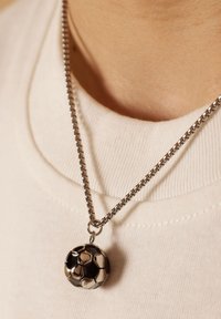 Metalen ketting met een kleine, gedetailleerde zwart-wit voetbalsymbool die aan een lichtgekleurde shirt hangt.