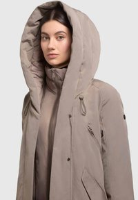 Béžový zateplený parka s veľkou kapucňou, predným zipsom a bočnými vreckami. Má hladkú textúru a minimalistický dizajn.