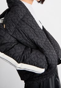 Kurtka puchowa w kolorze czarnym z tłoczonym wzorem logo. Posiada białe akcenty na rękawach oraz ściągacz na dole. Teksturowany materiał.