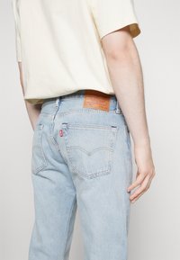 Jeans de denim azul claro con un diseño clásico de cinco bolsillos, detalles de costura en los bolsillos traseros y una etiqueta de marca de cuero marrón en la cinturilla.