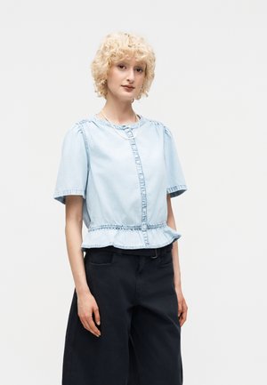 Νεαρό άτομο με ξανθά σγουρά μαλλιά που φοράει ανοιχτό μπλε κοντομάνικο τοπ με κουμπιά και βολάν στη μέση και μαύρο παντελόνι με φαρδιά πόδια.