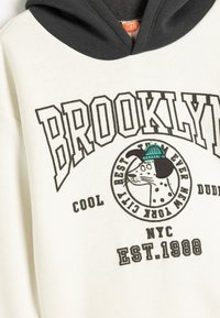 Kermakuvioinen huppari, jossa on tumma harmaa huppu. Koristeena on rohkea "BROOKLYN" -teksti ja graafinen kuva koirasta, jolla on vihreä hattu. Puuvillamateriaalia.