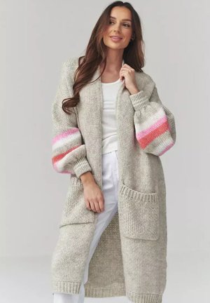 Kobieta w długim beżowym dzianinowym kardiganie z różowymi i czerwonymi paskami na bufiastych rękawach, białej bluzce i białych spodniach, stojąca na tle szarego tła.