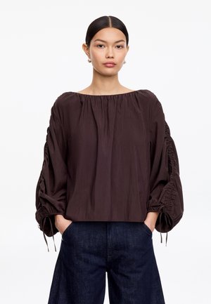 Mujer con cabello lacio y oscuro, vestida con una blusa marrón oscuro suelta con mangas anchas fruncidas y pantalones anchos azul oscuro, con las manos en los bolsillos.