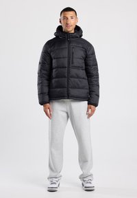 Black puffer jacket with a hood, featuring horizontal stitching, zippered chest pocket, worn over gray sweatpants and sneakers.