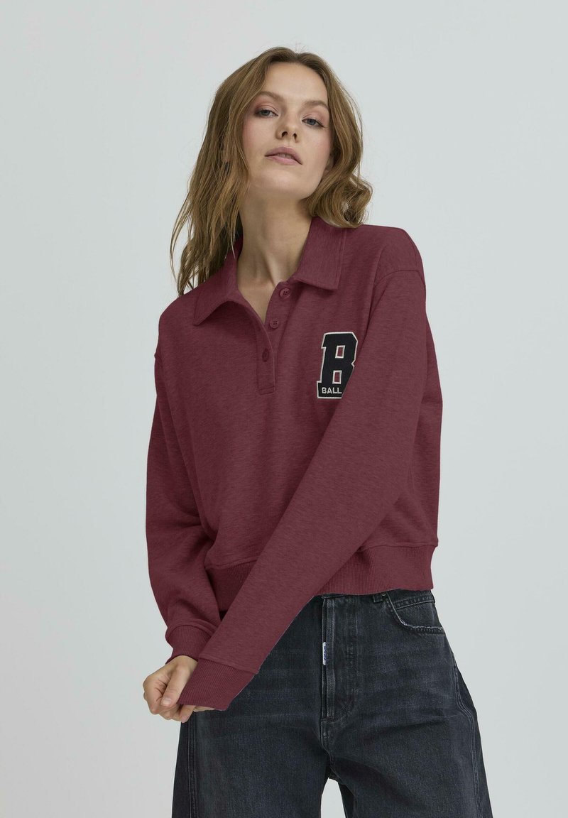 Bordó kapucnis pulóver, galléros nyakkal, három gombos gombolással és hímezett "B BALL" felirattal; sötét farmernadrággal párosítva.