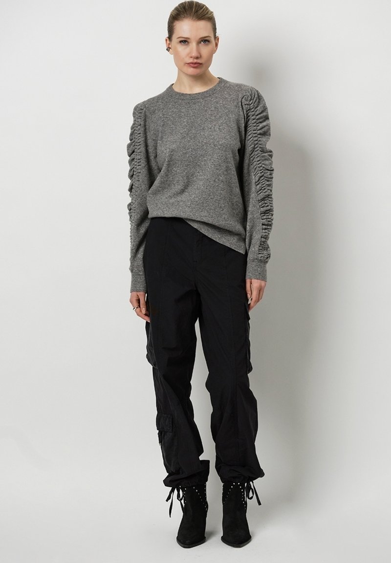 Grijze trui met textuur en gerimpelde mouwen, gecombineerd met zwarte cargo broeken en enkellaarsjes met studaccenten.