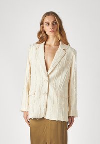 Blazer color crema con un diseño texturizado y deshilachado, dos botones y bolsillos frontales, con solapa de muesca y corte holgado.