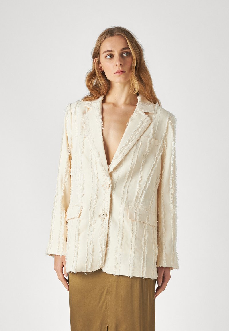 Blazer color crema con un diseño texturizado y deshilachado, dos botones y bolsillos frontales, con solapa de muesca y corte holgado.