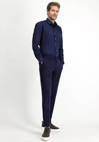 Granatowa koszula z długim rękawem na guziki, połączona z dopasowanymi wąskimi spodniami oraz brązowymi wsuwanymi trampkami. Gładka tkanina.