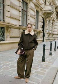 Chaqueta de lana marrón con un cuello de borreguito crema, pantalones anchos marrones, sosteniendo un pequeño bolso burdeos, de pie en una calle empedrada.