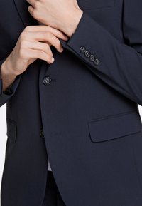 Giacca da abito su misura blu navy con rever a lancia, due tasche frontali e sei bottoni neri sul polsino. Tessuto dalla trama liscia.