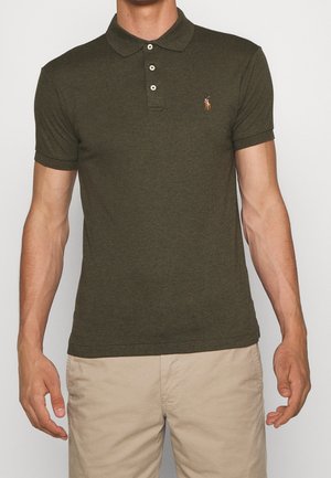 Mann trägt ein dunkelgrünes Poloshirt mit kurzen Ärmeln und drei Knöpfen sowie eine beige Hose, steht vor hellem Hintergrund.