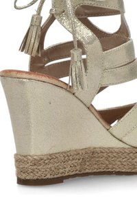 Sandalia cuña metálica dorada con punta abierta y diseño cortado. Presenta flecos decorativos y una cuña envuelta en yute.