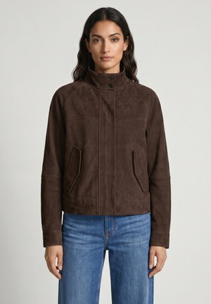Kvinde med langt mørkt hår iført en mørkebrun knappet jakke og blå jeans, stående mod en ensartet lys baggrund.