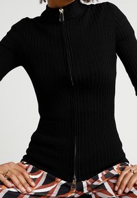 Pull en maille côtelée noir avec un col montant et une fermeture éclair sur toute la longueur, présentant un design ajusté et un tissu texturé. Assorti à un pantalon à motifs.