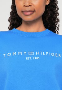 Modrá mikina vyrobená z bavlny s rebrom v oblasti krku. Na prednej strane je biele logo "TOMMY HILFIGER EST. 1985".
