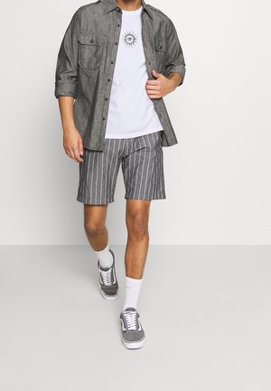 Chemise grise boutonnée, t-shirt blanc avec graphisme de soleil, short rayé gris et blanc, chaussettes blanches et baskets grises. Tenue décontractée présentée.