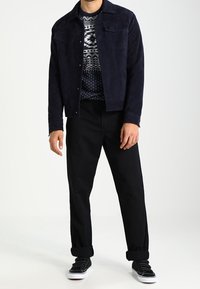 Marinblå mockajacka med två bröstfickor, mönstrad mörk tröja, svarta uppvikta byxor och svarta sneakers med vita sulor.