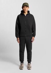 Zwarte zip-up hoodie met een capuchon, gecombineerd met bijpassende sweatpants. De outfit heeft een gladde textuur en elastische manchetten. Ggedragen met beige sneakers.