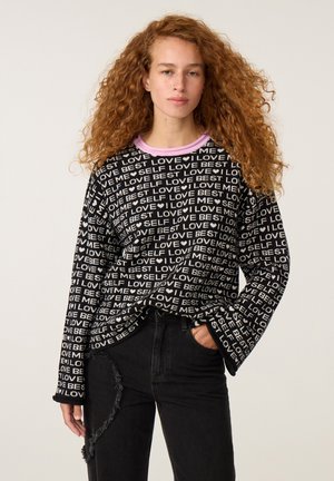 Vrouw met lang krullend haar, die zwarte spijkerbroek en een zwarte trui draagt met witte tekst die "LOVE ME SELF BEST" herhaalt en een roze kraagzoom.