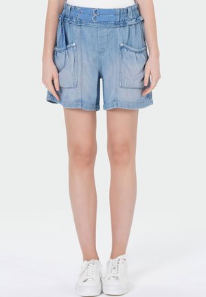 Short en jean bleu clair avec taille élastique et ceinture à double boucle, grandes poches avant appliquées, porté avec des baskets blanches.