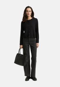 Frau mit langen, dunklen Haaren steht da und trägt eine schwarze, knöpfbare Strickjacke, dunkelgraue, gerade Jeans, schwarze Stiefel und hält eine schwarze Handtasche in ihrer linken Hand.