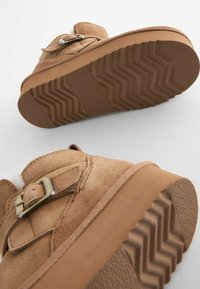 Scarpe in suede marrone con suola in gomma testurizzata, dotate di una cinghia con fibbia metallica e un leggero rivestimento in pelliccia vicino alla caviglia.