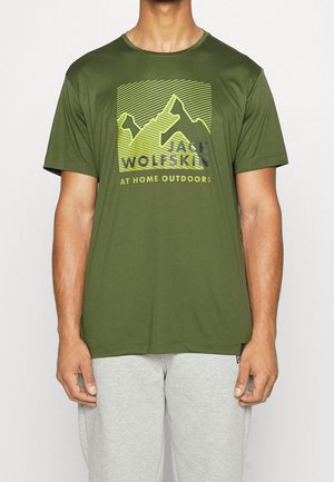 Olivově zelené triko s krátkým rukávem s horským grafikou v žlutém a šedém textu "Jack Wolfskin AT HOME OUTDOORS" na přední straně. Hladká textura.