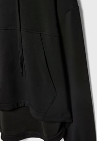 Sweat à capuche noir avec poche ventrale, coutures latérales et cordon sur fond uni.