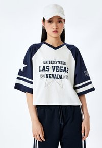 Camiseta de manga corta y escote en V en color crema y azul marino con gráfico de "LAS VEGAS", diseño de estrellas y rayas en las mangas. Combinada con pantalones azul marino.