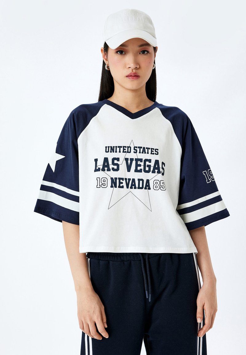 Camiseta de manga corta y escote en V en color crema y azul marino con gráfico de "LAS VEGAS", diseño de estrellas y rayas en las mangas. Combinada con pantalones azul marino.