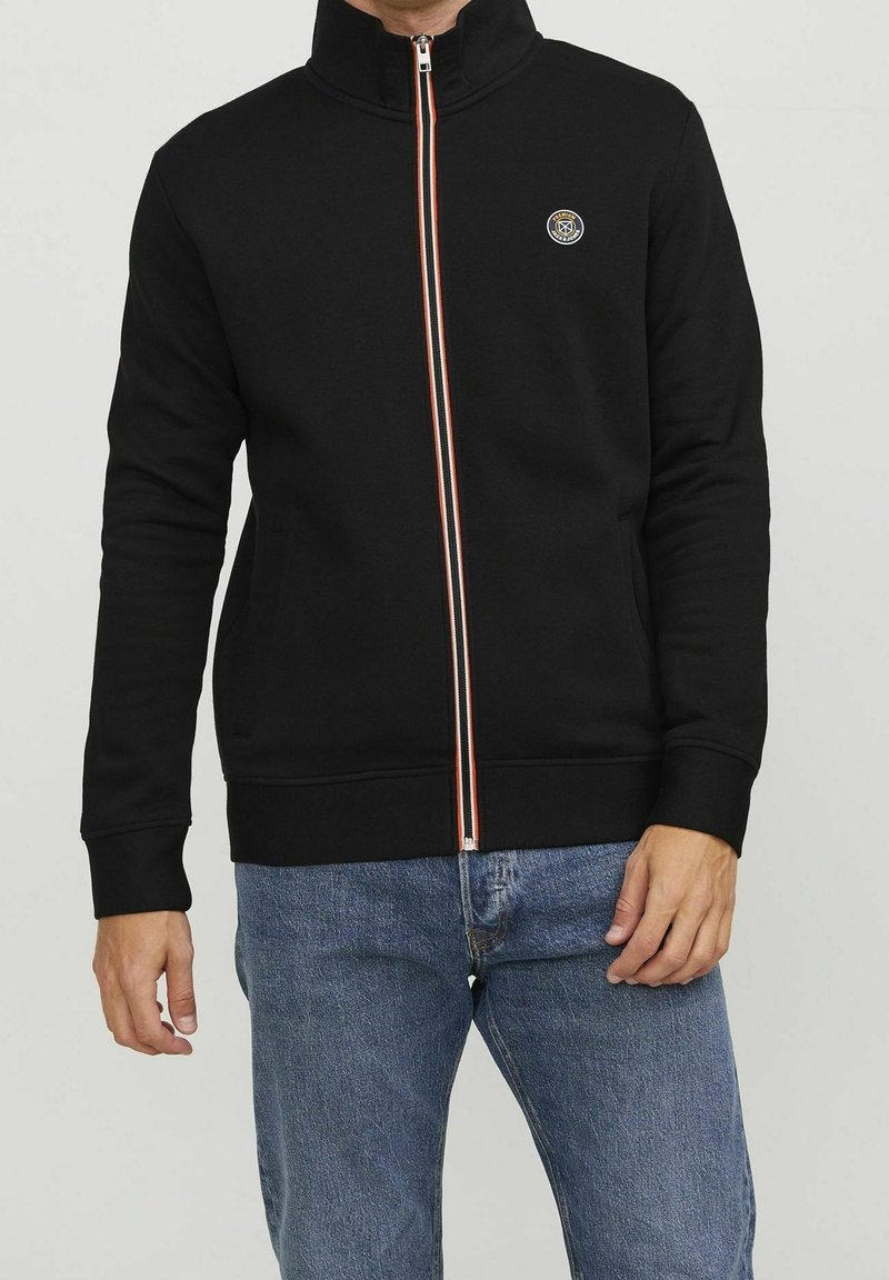Homme portant une veste noire zippée avec un petit logo rond sur la poitrine et un jean bleu, debout devant un fond clair uni.
