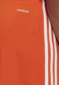 Shorts de sport orange avec des bandes blanches et le logo "AEROREADY". Fabriqués en tissu léger, avec une texture lisse sans poches.